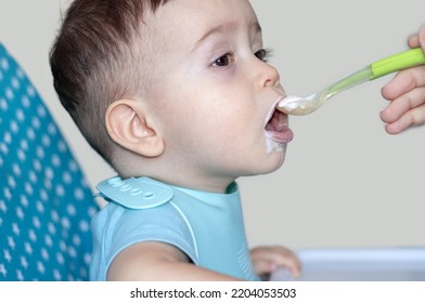 Mother Feeding Baby Boy With Mashed Food Puree Yogurt Healthy Diversification.toddler In High Chair Side View Green Silicone Spoon.wide Open Mouth Hungry Boy.wide Banner Child With Spoon In Hand.dirty