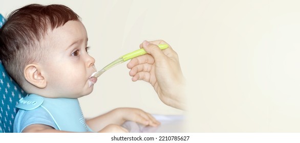 Mother Feeding Baby Boy With Mashed Food Puree Yogurt Healthy Diversification.toddler In High Chair Side View Green Silicone Spoon.wide Open Mouth Hungry Boy.wide Banner Child With Spoon In Hand.dirty