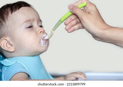 Mother Feeding Baby Boy With Mashed Food Puree Yogurt Healthy Diversification.toddler In High Chair Side View Green Silicone Spoon.wide Open Mouth Hungry Boy.wide Banner Child With Spoon In Hand.dirty