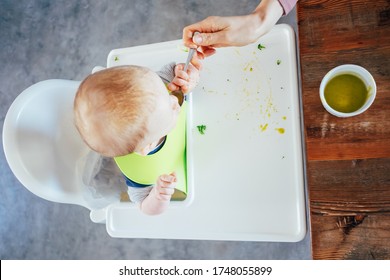 Mother feeding baby girl with puree. Above view of little child sitting on highchair and eating with help of her mother. Baby feeding concept - Shutterstock ID 1748055899