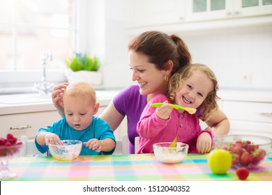 Mother Feeding Child Fruit And Yoghurt. Mom Feeds Kid In White Kitchen. Baby Boy And Girl Sitting In High Chair Eating Healthy Lunch Of Cereal And Milk. Nutrition, Healthy Breakfast For Toddler