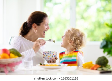 Mother Feeding Child Vegetables. Mom Feeds Kid In White Kitchen With Window. Baby Boy Sitting In High Chair Eating Healthy Lunch Of Steamed Carrot And Broccoli. Nutrition, Vegetarian Diet For Toddler