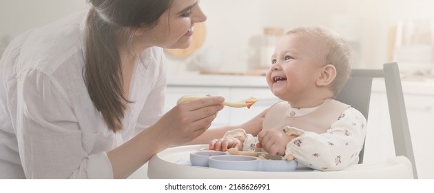 Mother Feeding Her Cute Little Baby In Kitchen. Banner Design