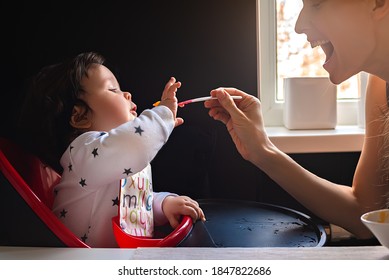 Mother feeding her little baby with a spoon in the kitchen on a black background.