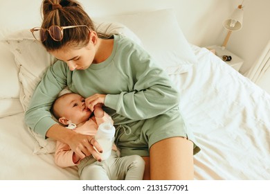 Mother Feeding Her Newborn Baby At Home. High Angle View Of A New Mom Holding A Bottle Of Formula Milk While Lying Next To Her Baby On The Bed. Single Mom Caring For Her Infant Child.