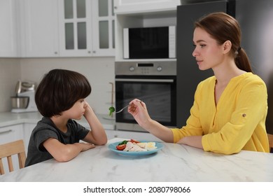 Mother Feeding Her Son In Kitchen. Little Boy Refusing To Eat Dinner