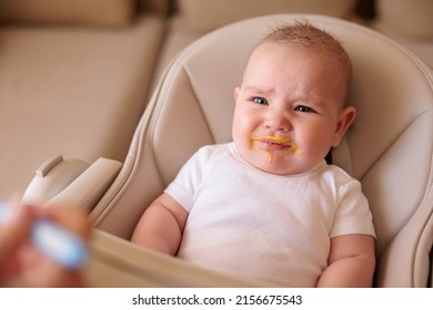 Mother feeding porridge to baby boy using spoon, introducing first solid food meal, angry baby sitting in high chair refusing food and crying