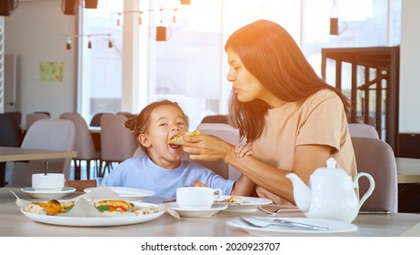 Mother Feeds Kid Girl In Restaurant. Asian Brunette Mom Holds Pizza Slice And Toddler Daughter Eats Sitting At Wooden Table With Tea In Local Cafe, Sunlight