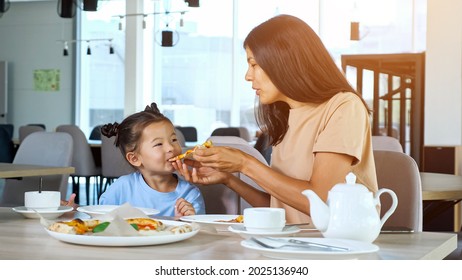 Mother Feeds Kid Girl In Restaurant. Asian Brunette Mom Holds Pizza Slice And Toddler Daughter Eats Sitting At Wooden Table With Tea In Local Cafe Close View