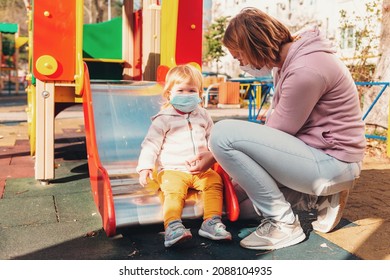 A Mother And Fer Little Child In Medicals Masks Sitting On A Children's Slide On Playground. Virus Protection In Public Places. Concept Of Covid-19 Pandemic.