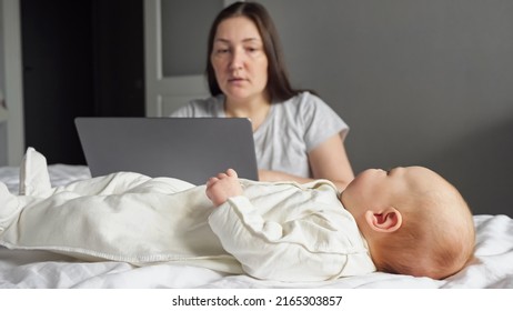 Mother Freelancer Looks After Baby Girl While Working On Laptop In Bedroom. Newborn Child Wiggles Legs And Arms On Parent Bed Distracting Mom From Work