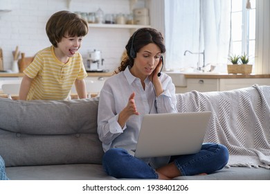 Mother Freelancer Talking On Video Call, Remote Working On Laptop From Home Office With Kid During Lockdown. Children Make Noise Disturb Woman At Work, Shows Tongue Standing Behind Couch. Distance Job