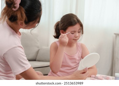 Mother gently helps daughter with makeup. The daughter holds a mirror, concentrating on her reflection. Beauty tools spread across the table, creating a tender mother-daughter bonding moment. - Powered by Shutterstock