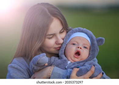 A mother gently holds her newborn son in her arms, looking lovingly at his facial features. A baby dressed in a bear costume yawns sweetly.