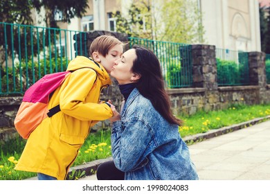 Mother gently kisses her son before school. Mom accompanies her child to school. Mother and pupil going to school in first class with schoolbag. Mother leads a little school boy in first grade.