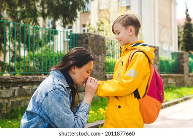Mother gently kisses her son before school. Mom accompanies her child to school. Mother and pupil going to school in first class with schoolbag. Mother leads a little school boy in first grade.