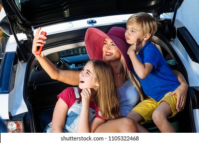 Mother, Girl And Boy Are Loading Multicolored Suitcases In The Trunk Of Car. Mom Makes Selfie On A Cell Phone With Her Children And Ready For Travel.