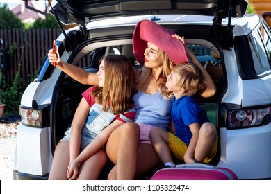 Mother, Girl And Boy Are Loading Multicolored Suitcases In The Trunk Of Car. Mom Makes Selfie On A Cell Phone With Her Children And Ready For Travel.