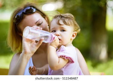 Mother Gives To Her Nice Baby Girl A Water From Bottle In Park. One-year-old Child In Pink Wants To Drink And Eat. Cut Toddler With Mom On A Walk. Beautiful Family With Kid On Blurry Background.