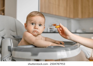 mother hand feeding a baby from a spoon. Mom giving a 9 month baby porridge. Meal time. Little boy eats sitting on baby's feeding chair. - Powered by Shutterstock