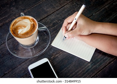 Mother Hand Holding Child Hand Writing On Notebook Over Wooden Table With Coffee Cup And Cellphone