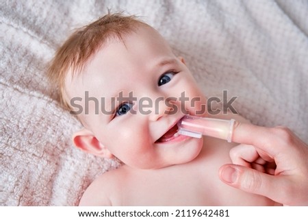 Mother hands brushing teeth with a finger brush of a happy infant baby. Mom doing oral hygiene to a smiling toddler kid, six to seven months old