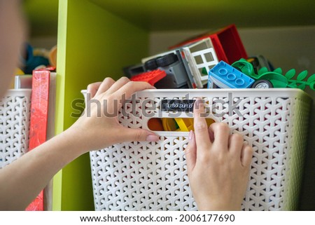 Mother hands sticking printing label sticker with name title of toy for comfortable storage and sorting in plastic case box. Woman arms use marker method for space organizing at children's room
