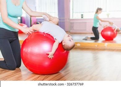 Mother With Happy Baby Doing Exercises With Red Gymnastic Ball At Fitness Class. Concept Of Caring For The Baby's Health.