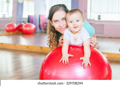 Mother With Happy Baby Doing Exercises With Red Gymnastic Ball At Fitness Class. Concept Of Caring For The Baby's Health.