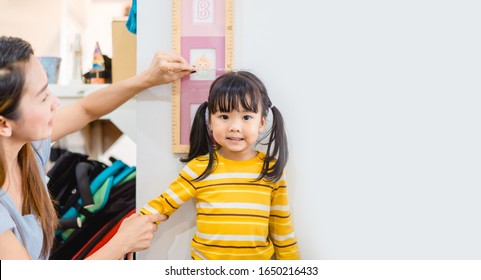 Mother Height Measure Her Daughter Near White Wall At Home.2.6 Years Old Girl Kid Standing And Looking At Above Using Hand Measuring Her Height.Growth Hormones, Child Development, Calcium Concept.