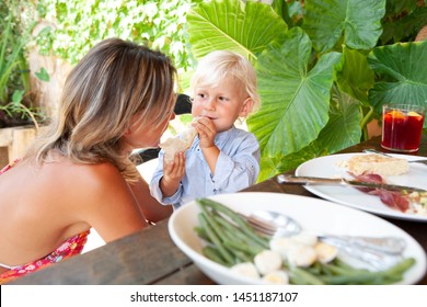 Mother Helping Child Son To Eat Lunch, Sitting At Family Dining Table In Lush Exotic Holiday Villa Garden Outdoors. Family On Sunny Summer Vacation Hotel Eating Together, Travel Leisure Lifestyle.