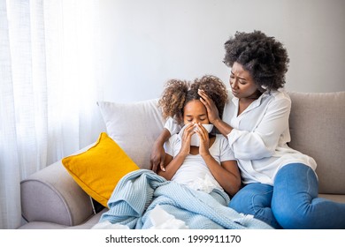 Mother helping daughter blowing her nose on the sofa at home. Ill African American girl blowing a nose at home while her mother is consoling her. Young mother feeling sad while looking at daughter - Powered by Shutterstock