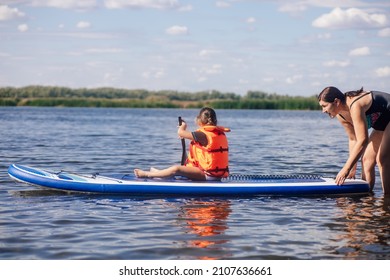 Mother helping little daughter to sup board with oar in hands on lake with green reeds and trees in background wearing vest life jacket. Active holidays. Inculcation of love for sports from childhood. - Shutterstock ID 2107636661
