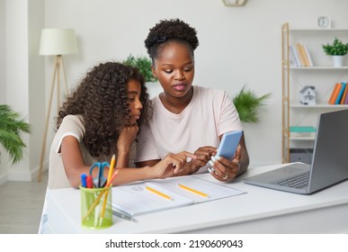 Mother helps her teenage daughter complete her school homework while using her smartphone. African American family sitting in front of notebook and laptop and looking at mobile phone screen together - Powered by Shutterstock