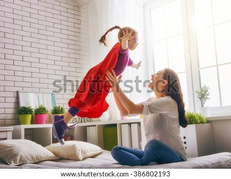 Mother and her child girl playing together. Girl in an Superman's costume. The child having fun and jumping on the bed.