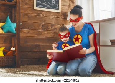 Mother And Her Child Playing Together. Girl And Mom In Superhero Costumes. Mum And Kid Having Fun, Smiling And Reading A Book. Family Holiday And Togetherness.