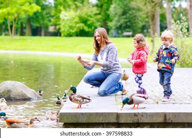 Mother And Her Children Feeding Ducks In Summer Park, Adorable Kid Boy And Girl, Siblings Having Fun Together