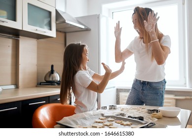 Mother And Her Cute Cheerful Daughter At Home Standing At Table In Kitchen Together Kid Giving High Five With Dirty Hands In Flour