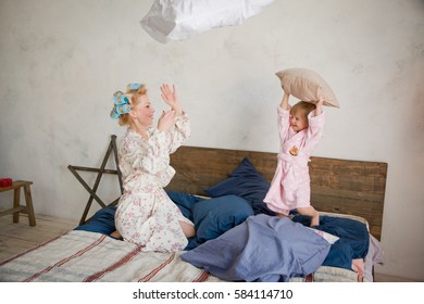 Mother With Her Daughter In A Pillow Fight On The Bed. They Are Happy And Having Fun. Play In The Morning Together. The Sleeping Girl's Clothes Having Fun. In Her Hair And In Robes At Home