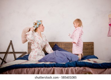 Mother With Her Daughter In A Pillow Fight On The Bed. They Are Happy And Having Fun. Play In The Morning Together. The Sleeping Girl's Clothes Having Fun. In Her Hair And In Robes At Home