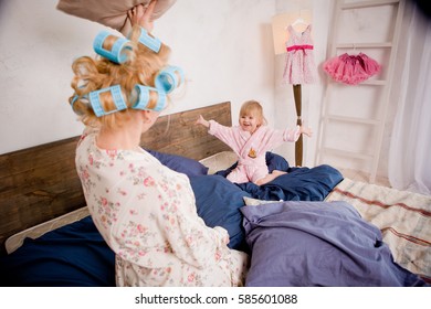 Mother With Her Daughter In A Pillow Fight On The Bed. They Are Happy And Having Fun. Play In The Morning Together. The Sleeping Girl's Clothes Having Fun. In Her Hair And In Robes At Home