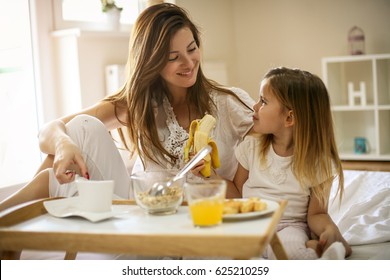 Mother with her little daughter having breakfast in the bed. Mother and her daughter enjoying a healthy breakfast together. Little girl eating banana. - Powered by Shutterstock