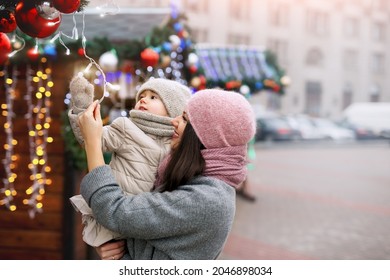 A Mother And Her Little Daughter Walk Through The City Streets And Christmas Markets Decorated With Garlands With Lights, Fir Branches And Christmas Balls. A Cold Frosty Day On New Year's Eve. Xmas