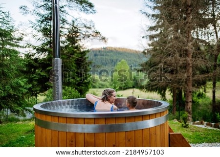 Mother with her little son enjoying bathing in wooden barrel hot tub in the terrace of the cottage. Wooden bathtub with a fireplace to burn wood and heat water. Rear view.