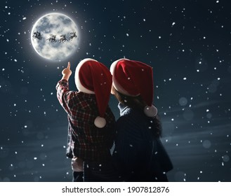Mother and her little son looking at Santa Claus with reindeers in sky on full moon night. Christmas holiday