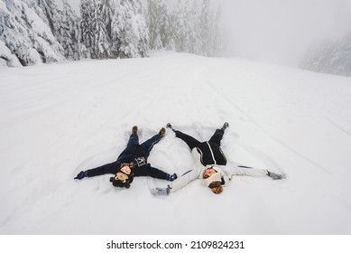 Mother And Her Son In Funny Hat Making Snow Angels Together Lying In Snowy Winter Forest. Happy Family Wearing Warm Winter Clothes Having Fun In Snow Covered Pine Forest. Outdoor Activities With Kids.