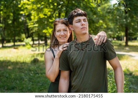 A mother and her son, who will soon be not a teenager but a young man, are chatting and having fun in the park. The boy is fooling around and the mother is proud of her son, smiles and hugs him
