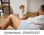 A mother and her toddler share a joyful moment at home. They are sitting on the wooden floor next to cozy, rustic decor. The scene captures a warm, loving family interaction.