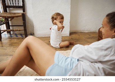 A mother and her toddler share a joyful moment at home. They are sitting on the wooden floor next to cozy, rustic decor. The scene captures a warm, loving family interaction. - Powered by Shutterstock