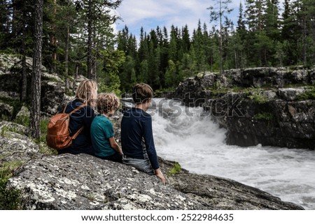 A mother and her two sons sit on a rock, captivated by the powerful Sognstupet Waterfall in Idre Dalarna surrounded by green trees and rushing water in the wilderness of Sweden